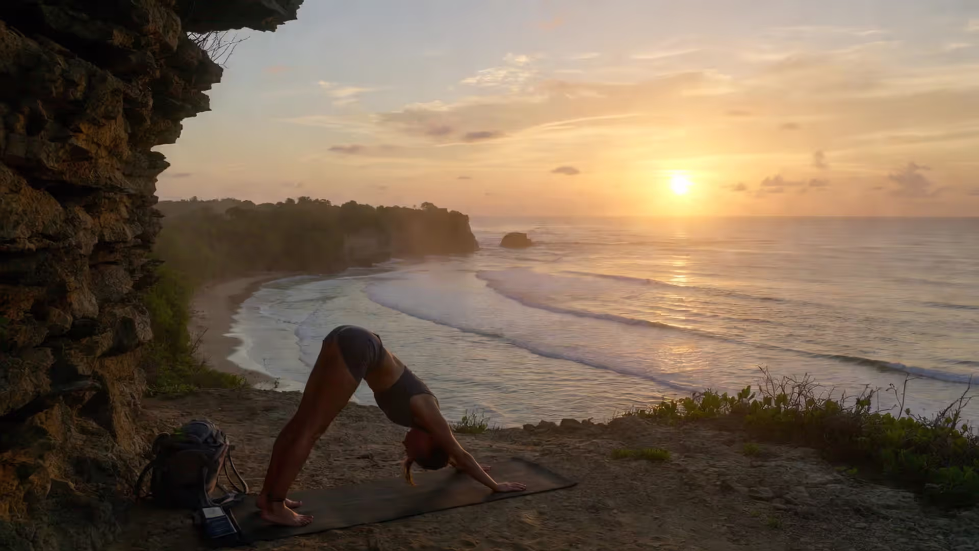 Traveler practicing yoga at sunrise with backpack beside mat overlooking ocean.